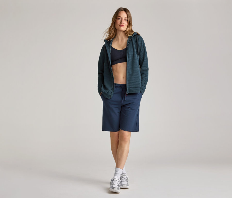 Femme portant un short et des chaussures de sport avec une veste à capuche bleu marine ouverte sur une brassière de sport.