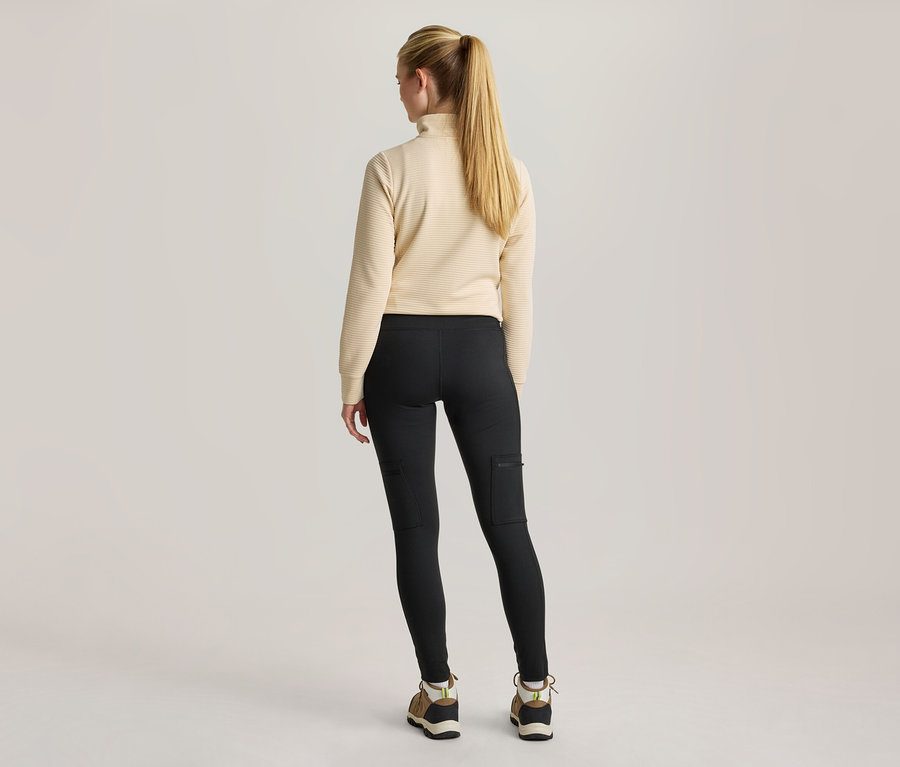 Une femme est debout, dos au spectateur, vêtue d'un haut côtelé à manches longues de couleur crème, d'un legging noir avec des poches latérales et de chaussures de randonnée.