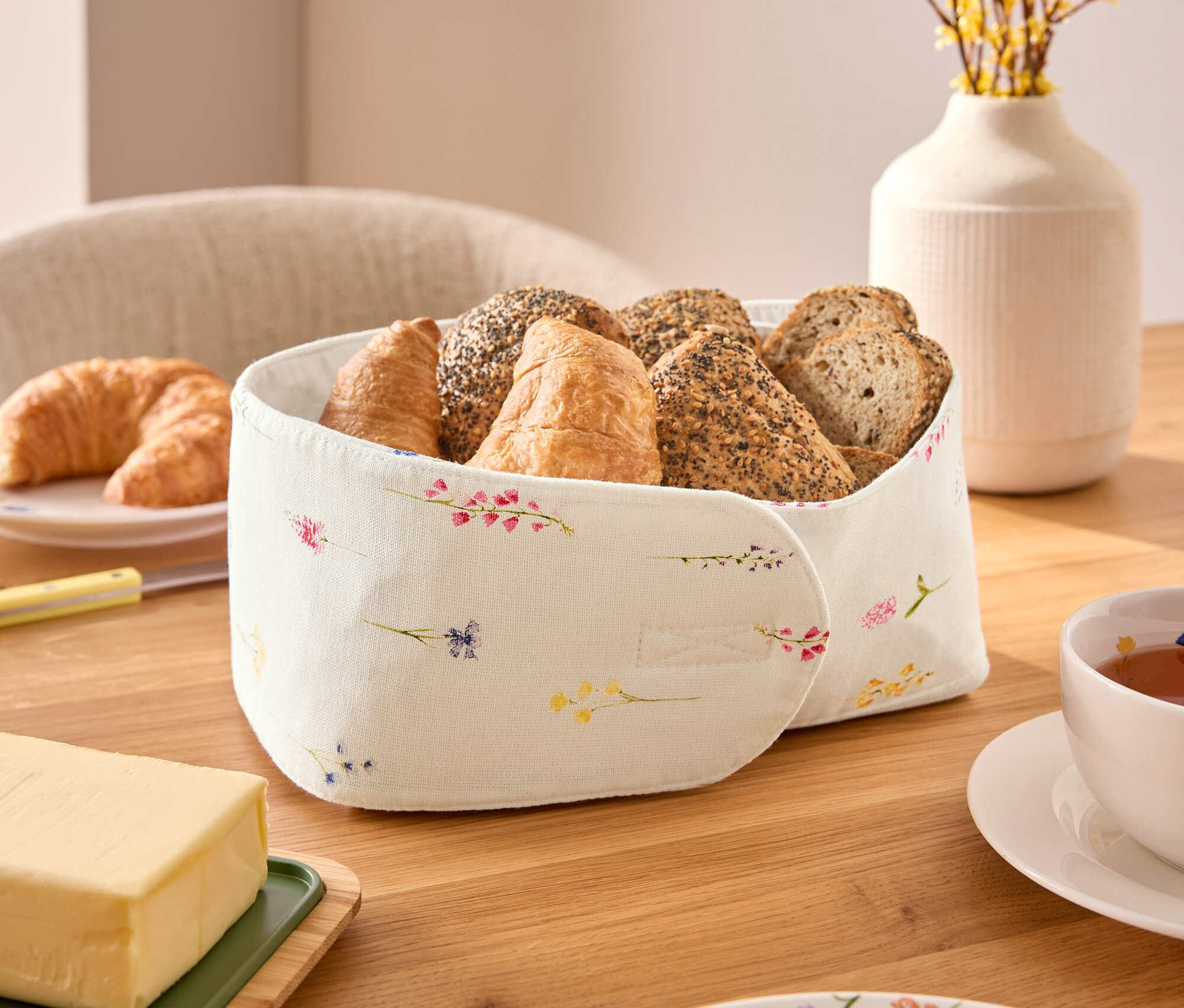 Sur une table en bois, une corbeille à pain contient diverses sortes de pain, dont des croissants et des petits pains, à côté d'une tasse de thé, d'une portion de beurre et d'une assiette à motifs floraux.