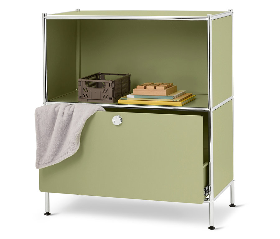 Une armoire verte avec un compartiment ouvert sur le dessus contenant une caisse marron, des livres et des blocs de bois. Un tissu gris est suspendu au-dessus du tiroir.