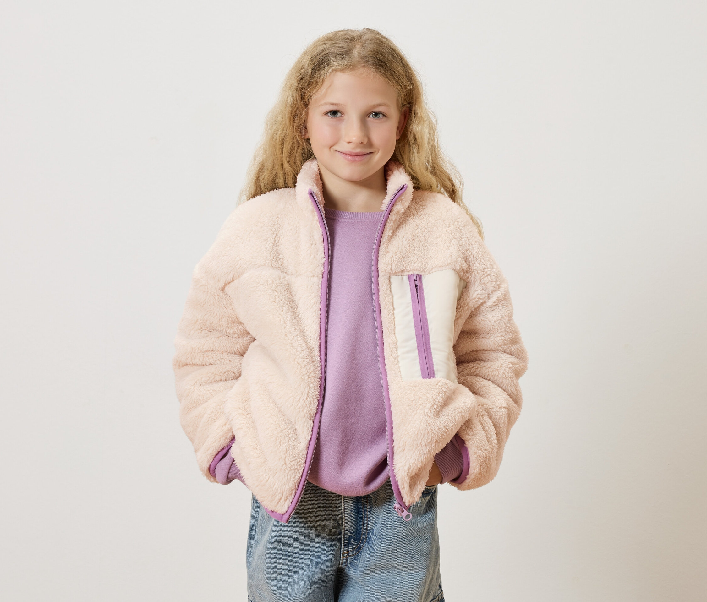 Une fille aux cheveux blonds bouclés porte une veste en teddy rose sur un pull violet et un jean bleu.