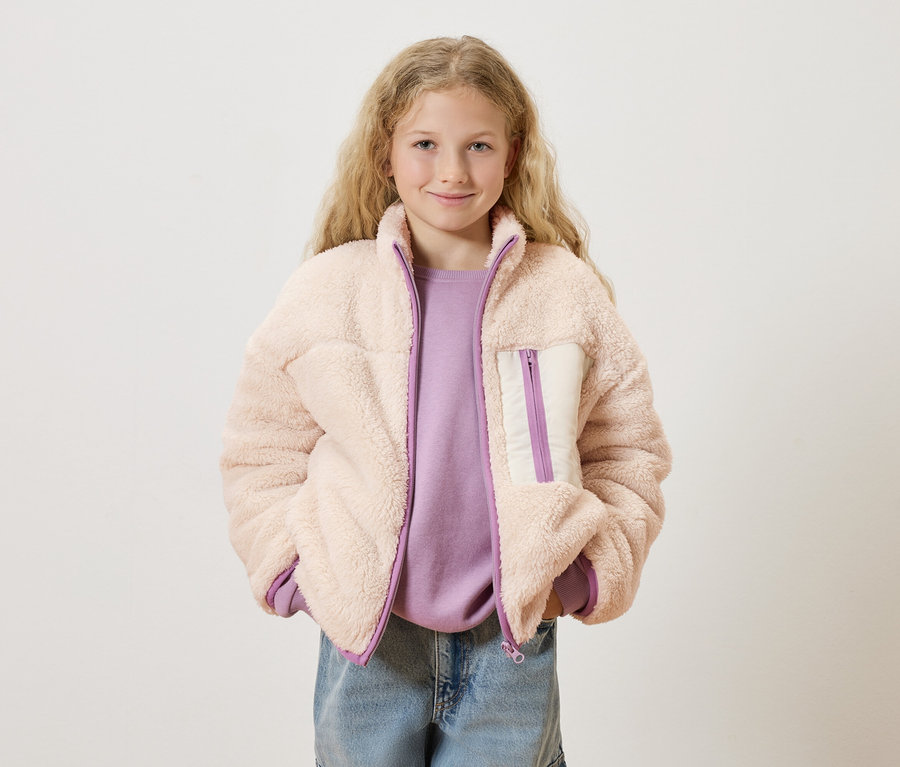 Une fille aux cheveux blonds bouclés porte une veste en teddy rose sur un pull violet et un jean bleu.