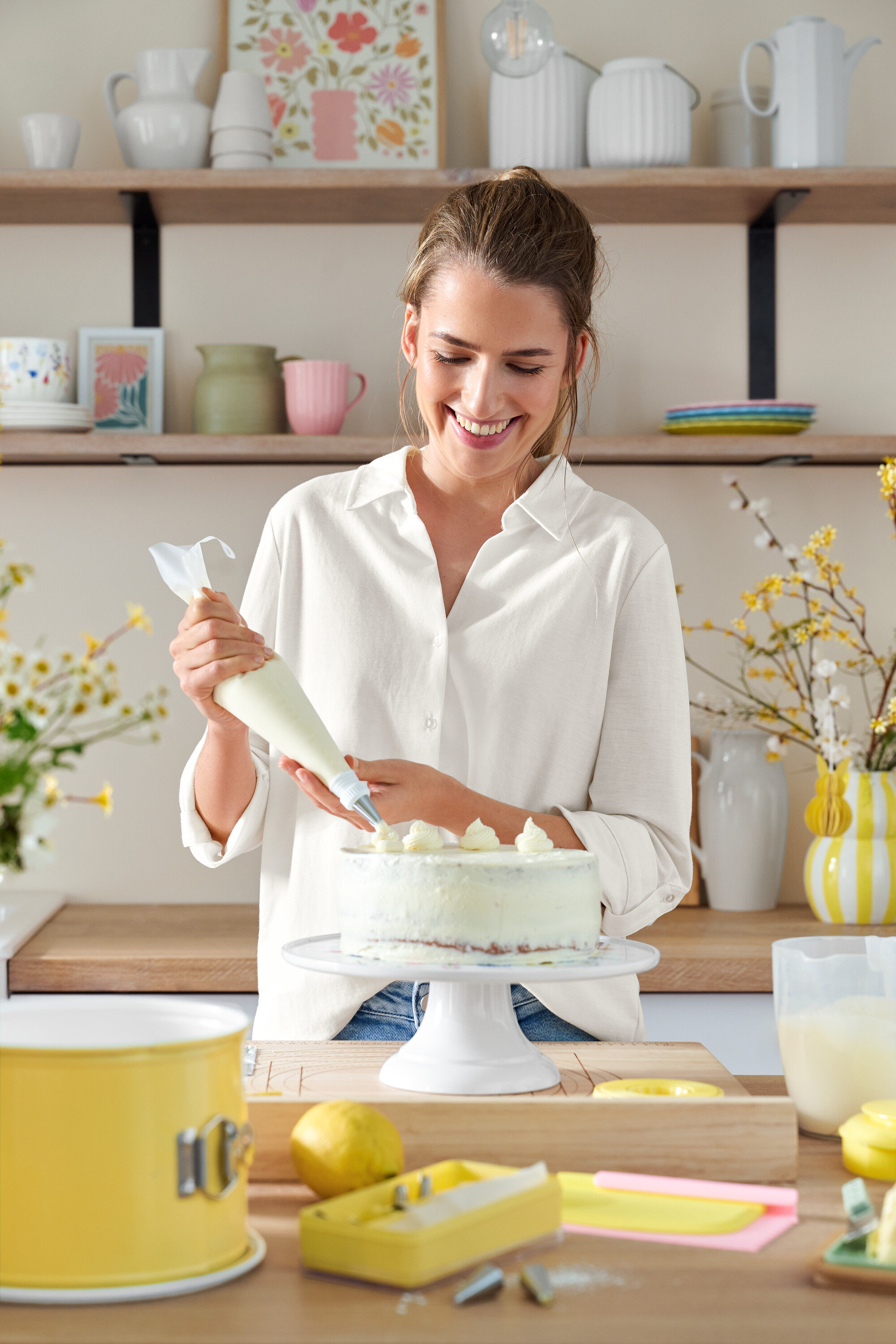 Une femme décore un gâteau avec une poche à douille. Des étagères avec des ustensiles de cuisine sont visibles en arrière-plan.