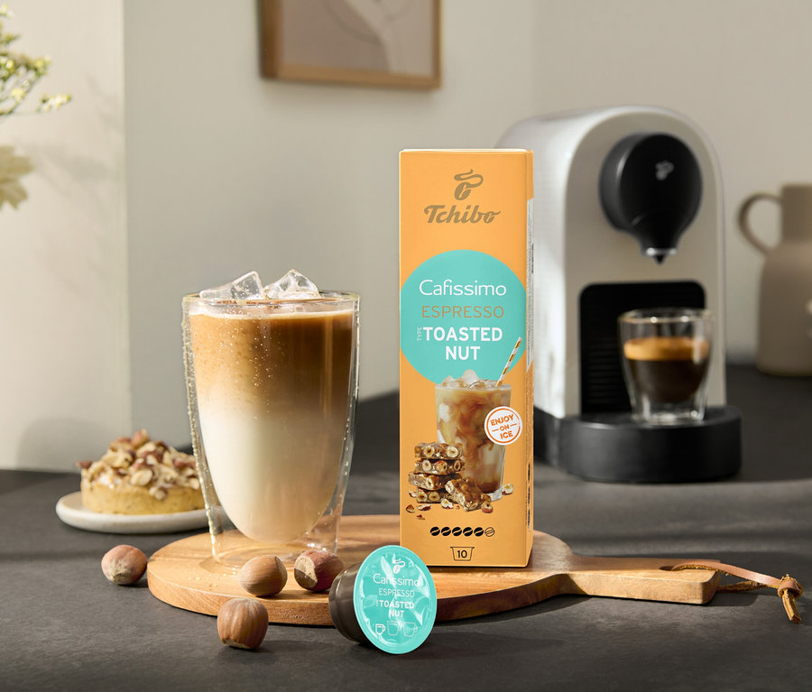 Un verre de café glacé est à côté d'un paquet de Cafissimo Flavoured Espresso – Toasted Nut et d'une machine à café Cafissimo pure+, white.