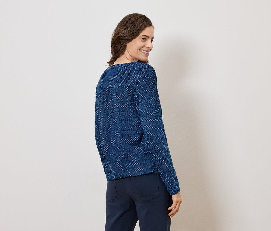 Femme portant un chemisier bleu à motifs et un pantalon foncé, vue de dos.