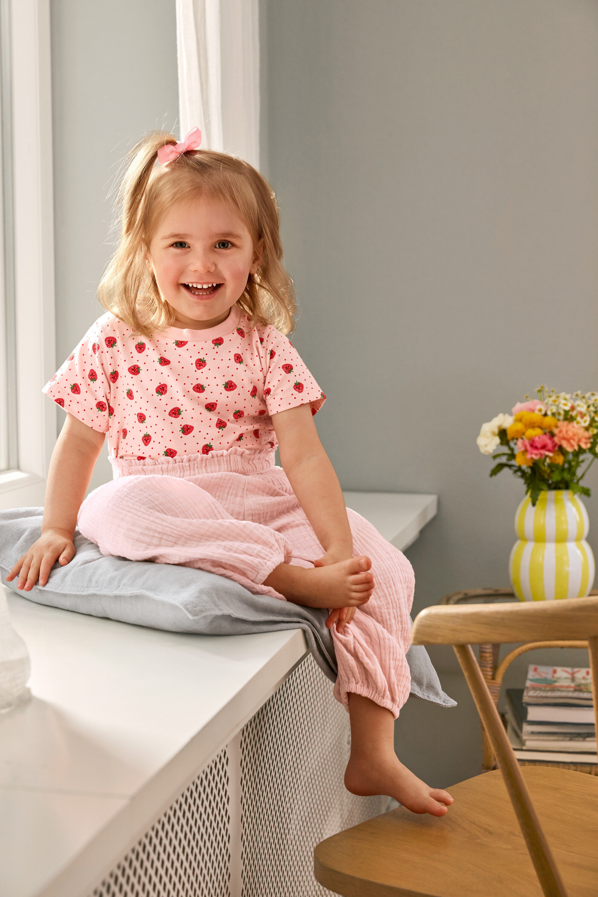 Une fillette souriante est assise sur un coussin en portant 2 Kinder-T-Shirts et un Kinder-Musselin-Hose. Un vase est posé sur une chaise.
