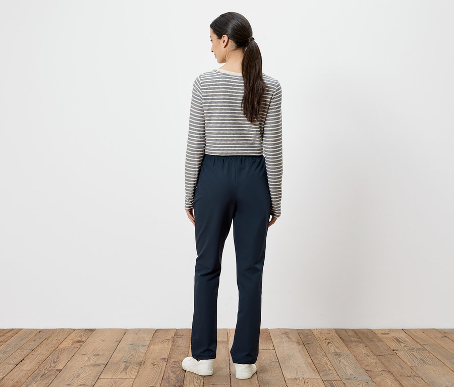 Une femme avec une queue de cheval se tient dos au spectateur, portant un t-shirt rayé à manches longues, un pantalon bleu marine et des baskets blanches.