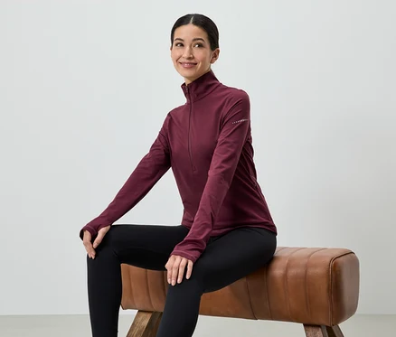 Une femme est assise sur un tabouret, portant un haut bordeaux à manches longues et un legging noir.