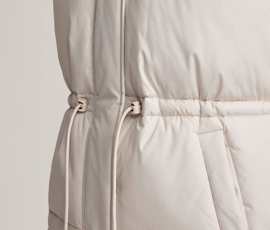Gros plan d'une doudoune sans manches beige clair avec un cordon de serrage à la taille et deux stoppeurs. Un haut à manches longues rayé vert et blanc est visible en dessous.