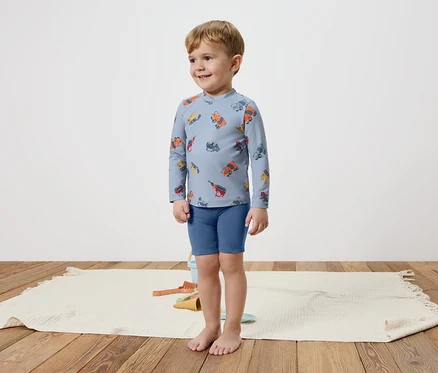 Un petit garçon se tient pieds nus sur un tapis, portant un haut bleu à manches longues avec des véhicules colorés et un short bleu.