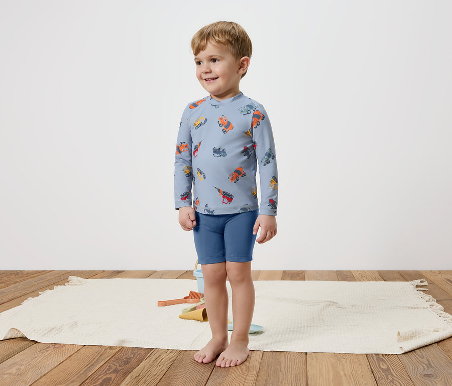 Un petit garçon se tient pieds nus sur un tapis, portant un haut bleu à manches longues avec des véhicules colorés et un short bleu.