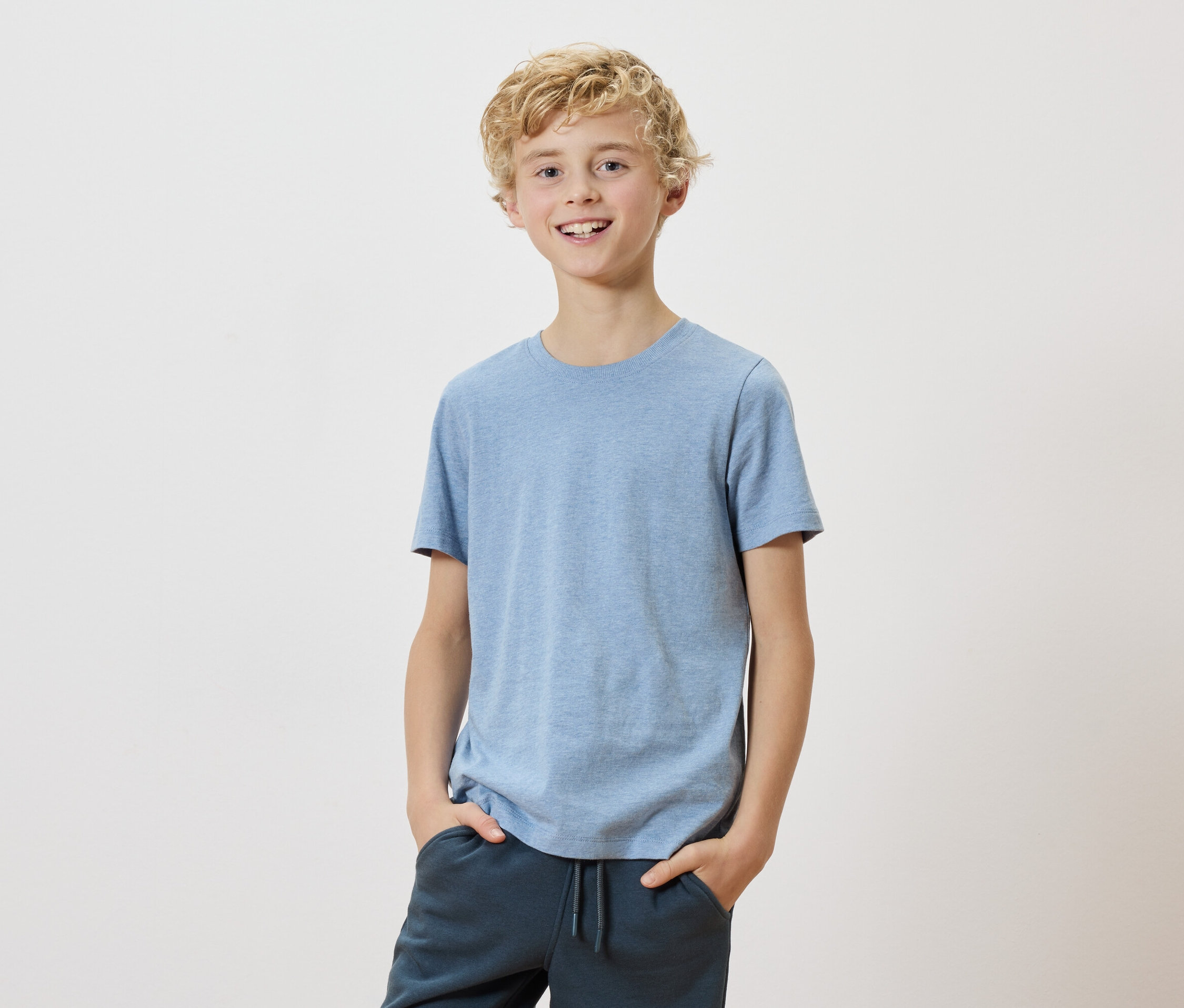 Un garçon blond avec un t-shirt bleu clair et un pantalon foncé se tient devant un fond blanc.