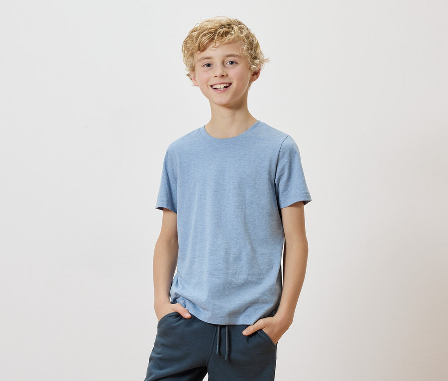 Un garçon blond avec un t-shirt bleu clair et un pantalon foncé se tient devant un fond blanc.