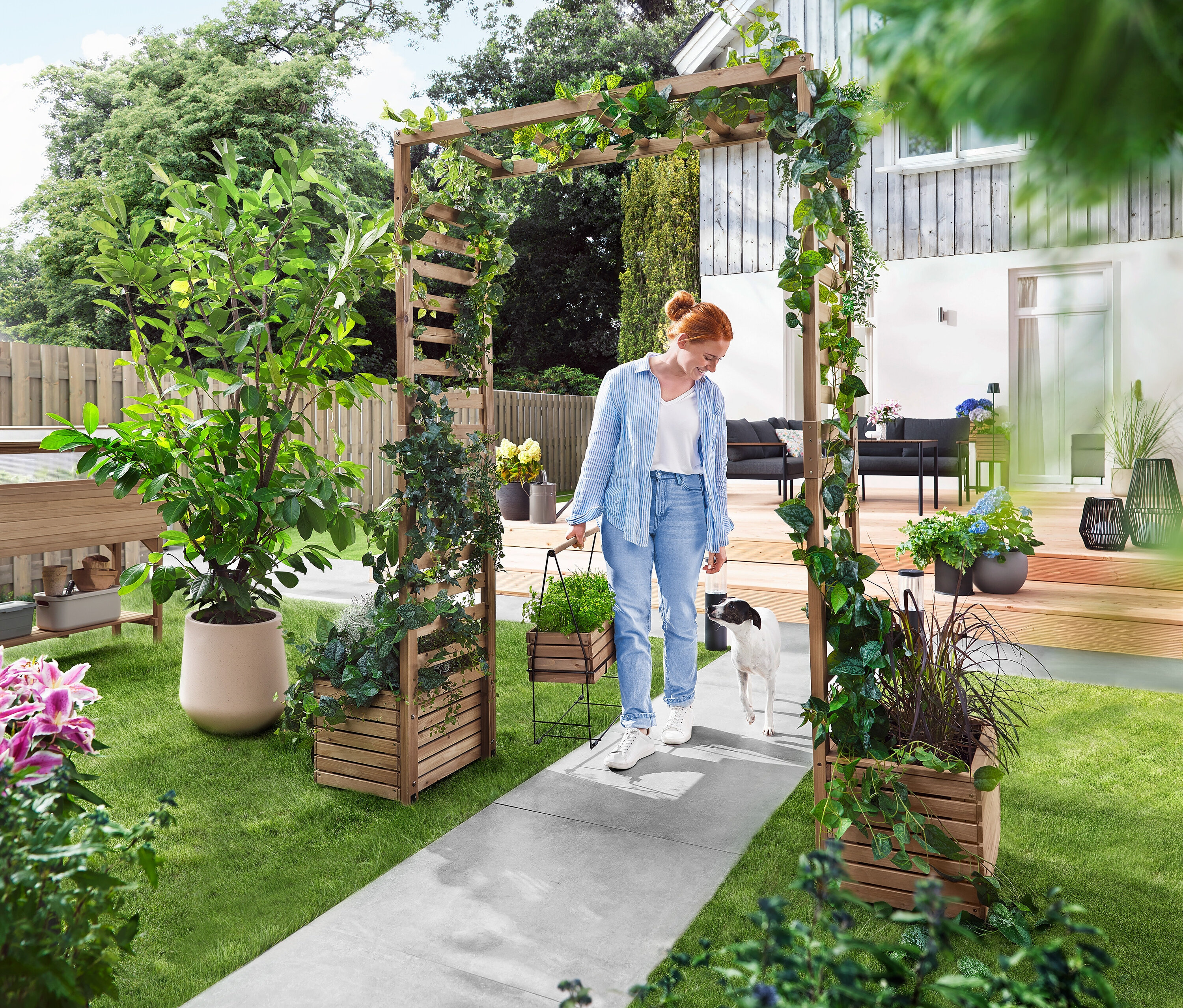 Une femme marche avec un chien à travers une arche de jardin recouverte de plantes. Elle porte un bac à plantes.