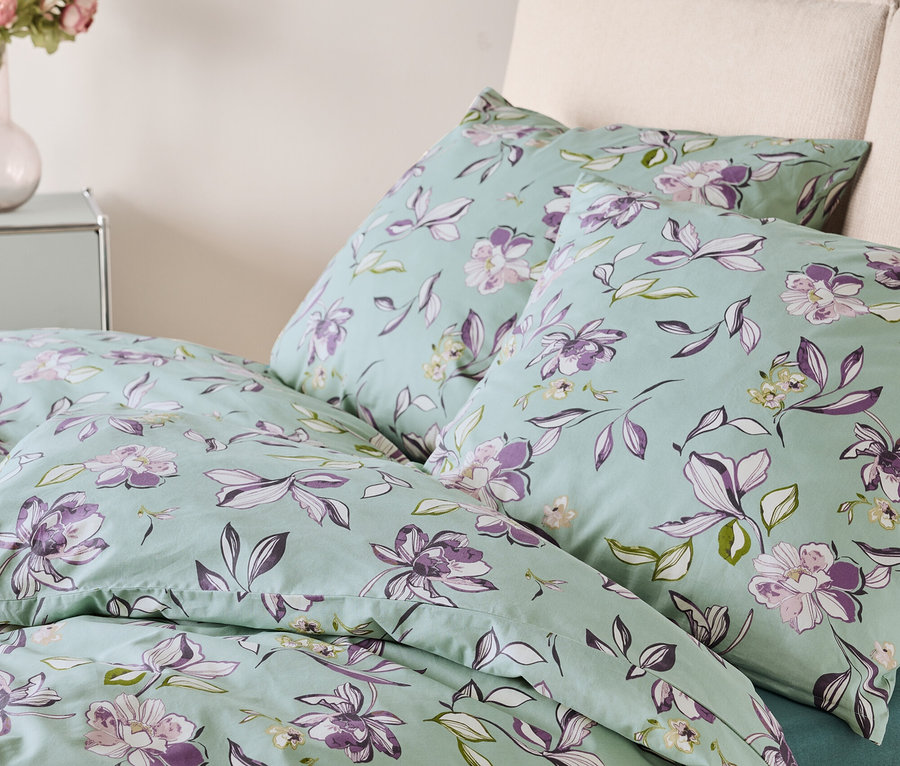 Lit avec des draps vert clair imprimés de fleurs violettes et blanches. Deux oreillers sont posés sur le lit.