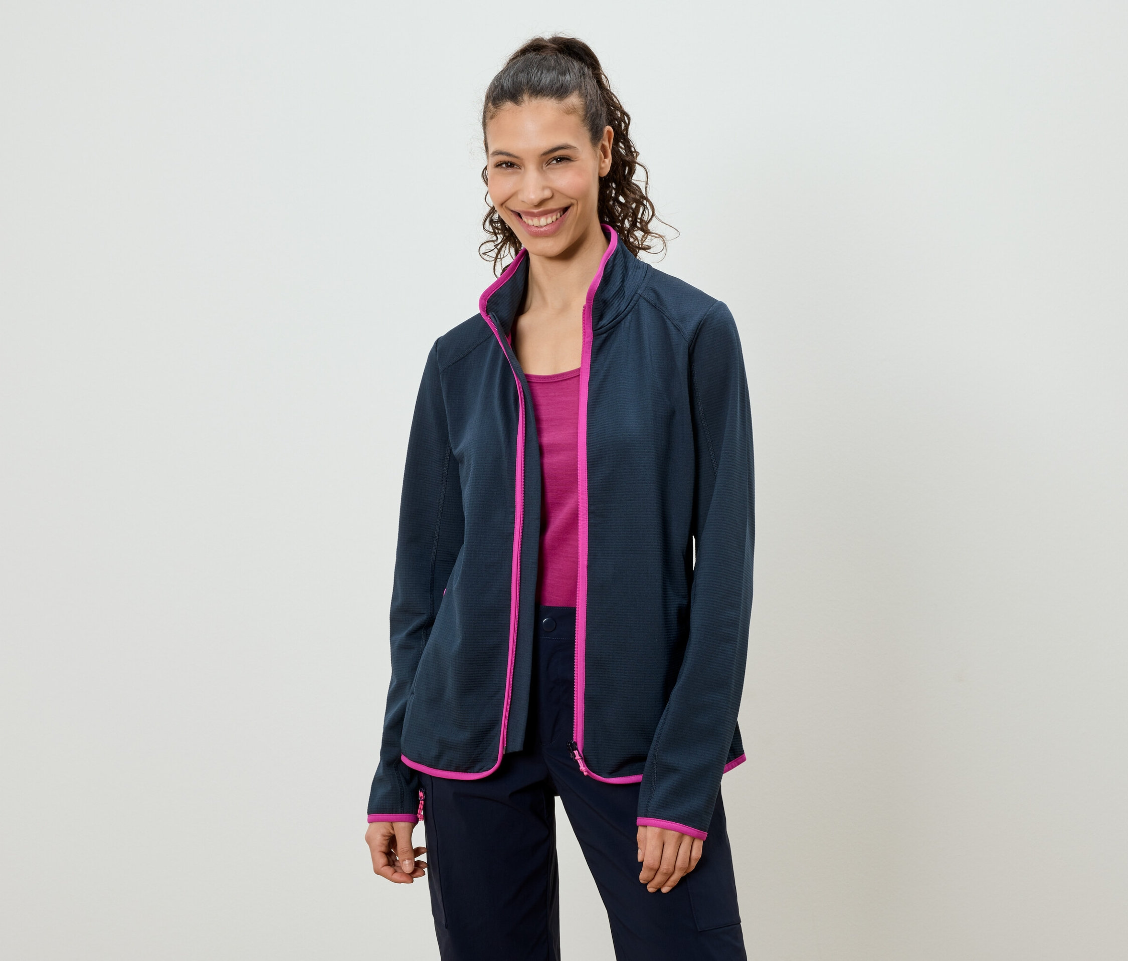 Une femme avec une queue de cheval porte une veste polaire bleu marine avec des touches de rose par-dessus un haut rose et un pantalon bleu marine.