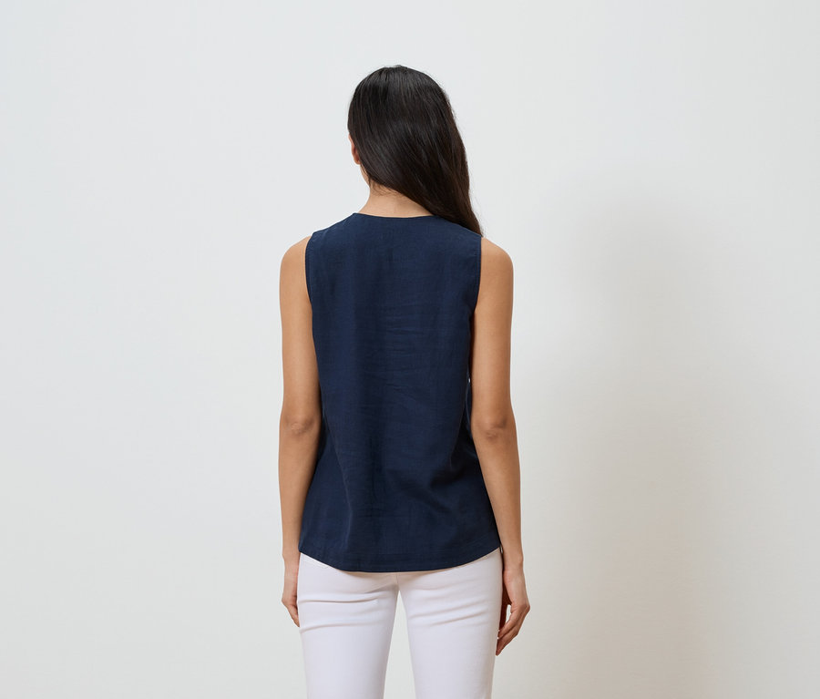 Femme aux cheveux foncés, vue de dos, portant un haut bleu marine sans manches et un pantalon blanc sur fond blanc.