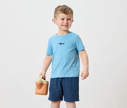 Un petit garçon aux cheveux blonds porte un t-shirt bleu clair avec un motif de requin et un short bleu marine. Il tient un arrosoir orange.