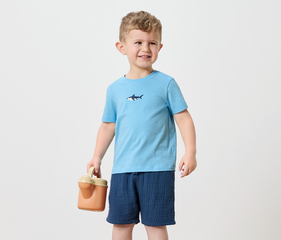 Un petit garçon aux cheveux blonds porte un t-shirt bleu clair avec un motif de requin et un short bleu marine. Il tient un arrosoir orange.