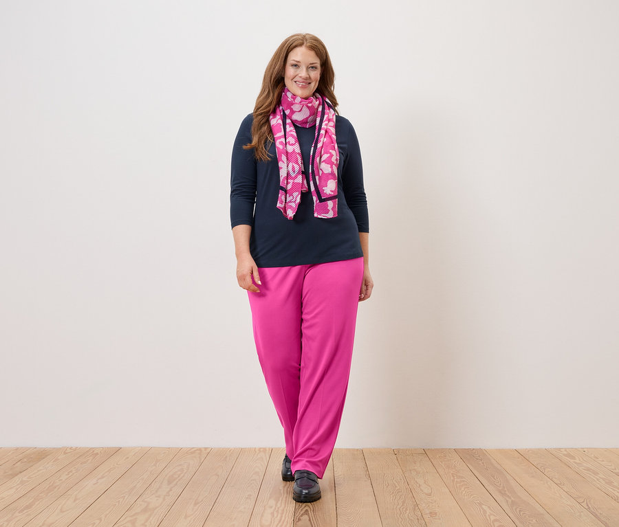 Une femme aux longs cheveux roux se tient devant un mur blanc, portant un chemisier bleu marine, un pantalon rose et un foulard rose à fleurs.