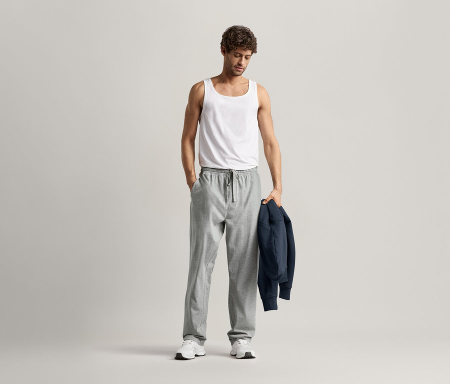 Homme portant un débardeur blanc et un pantalon relax gris, une main dans la poche et une veste bleu marine sur le bras.