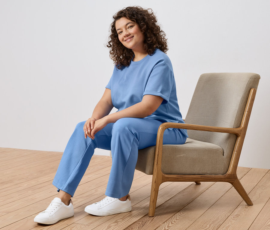 Une femme aux cheveux bouclés est assise dans un fauteuil, portant un t-shirt bleu clair, un pantalon et des baskets blanches.