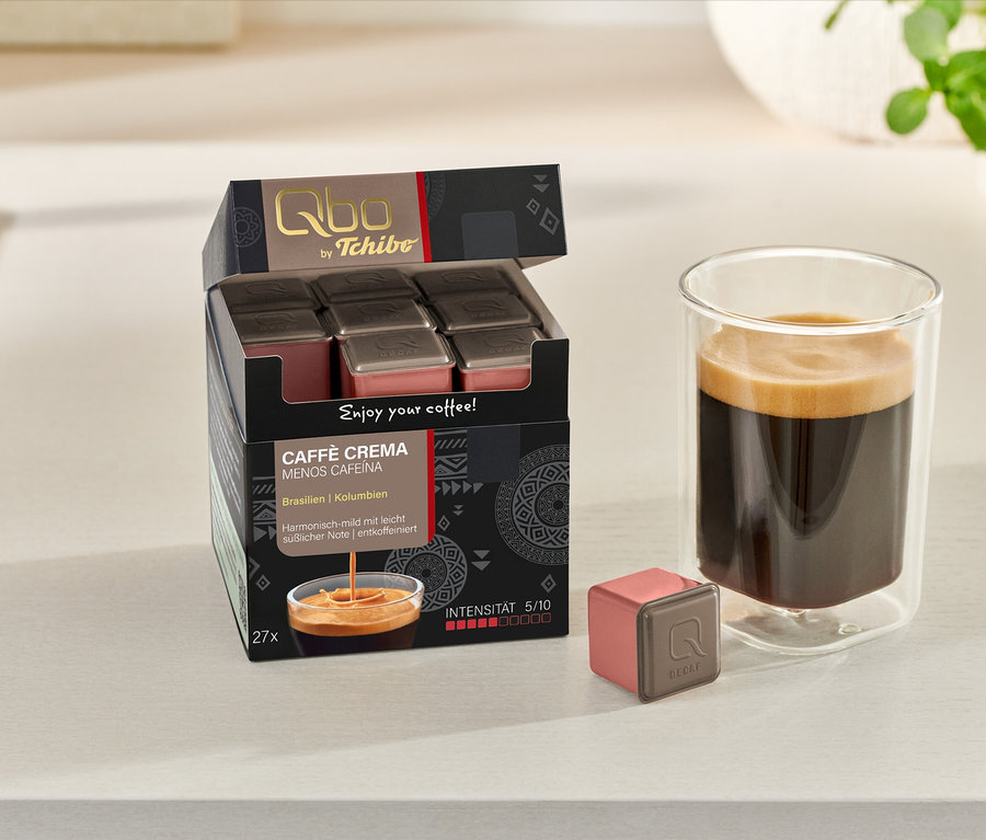 Un paquet ouvert de Qbo Caffè MENOS CAFEÍNA de 27 capsules, une capsule et un verre de café sont posés sur une surface claire.