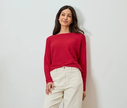 Femme appuyée contre un mur, portant un pull rouge et un pantalon beige.