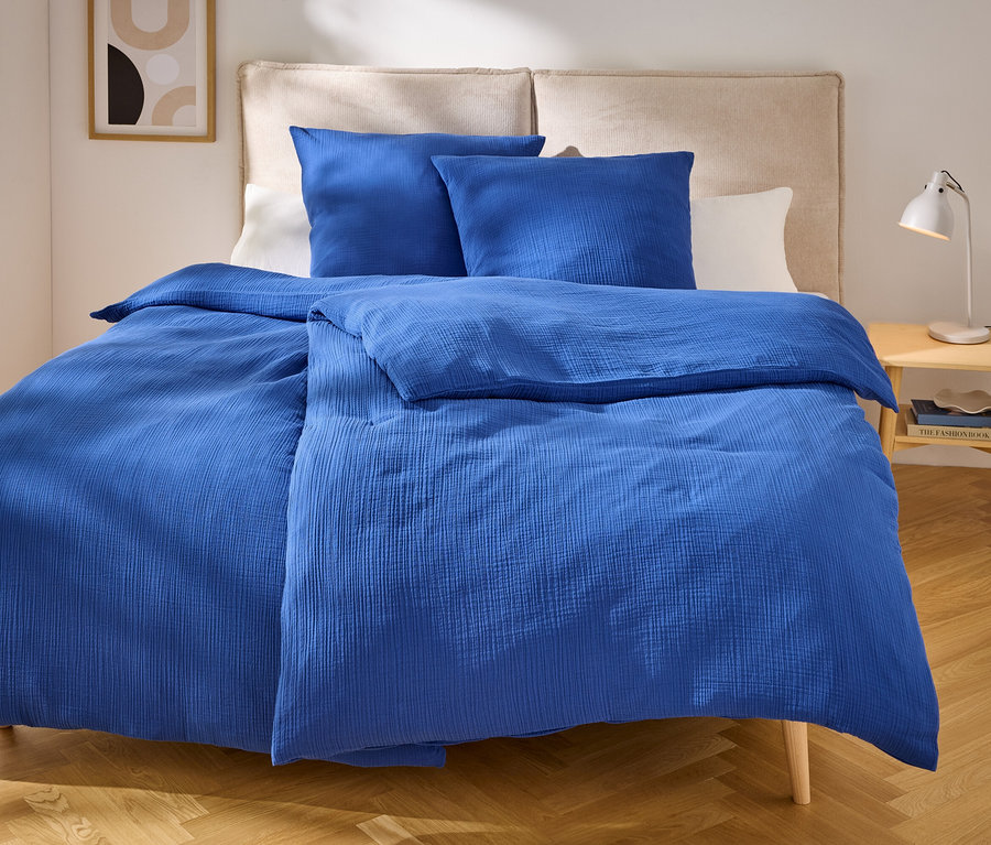 Literie bleue avec deux oreillers sur un lit avec une tête de lit beige. Une lampe est posée sur la table de chevet.