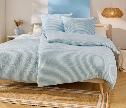 Lit avec des draps rayés bleu clair et blanc, deux oreillers et une couverture. Un tapis beige est placé sous le lit.