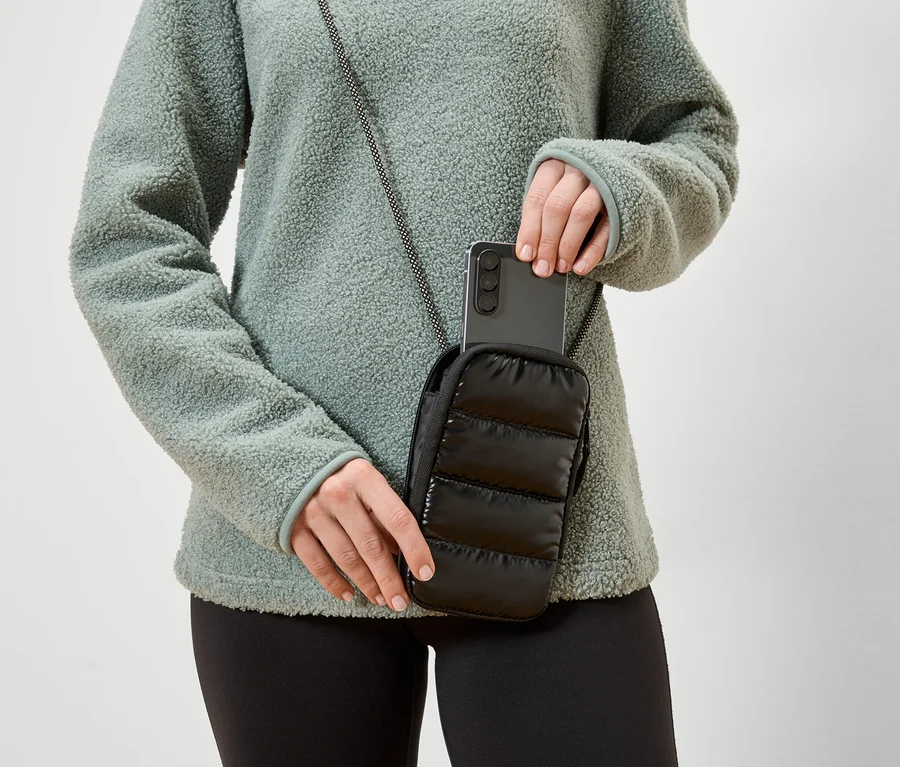 Une femme glisse un smartphone dans une pochette noire matelassée pour téléphone avec un cordon qu'elle porte autour du cou. Elle porte un haut en polaire vert.