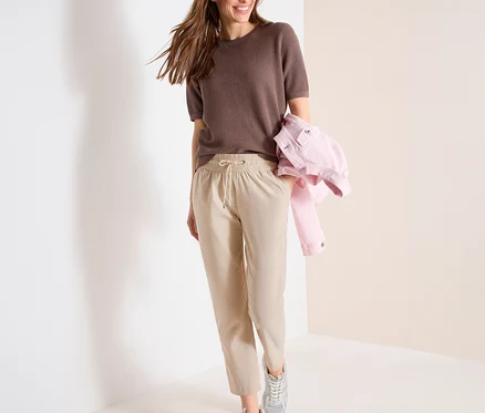 Femme souriante marchant, portant un haut marron texturé, un pantalon de jogging CECIL beige clair avec cordon de serrage et des baskets argentées, une veste rose sur le bras.