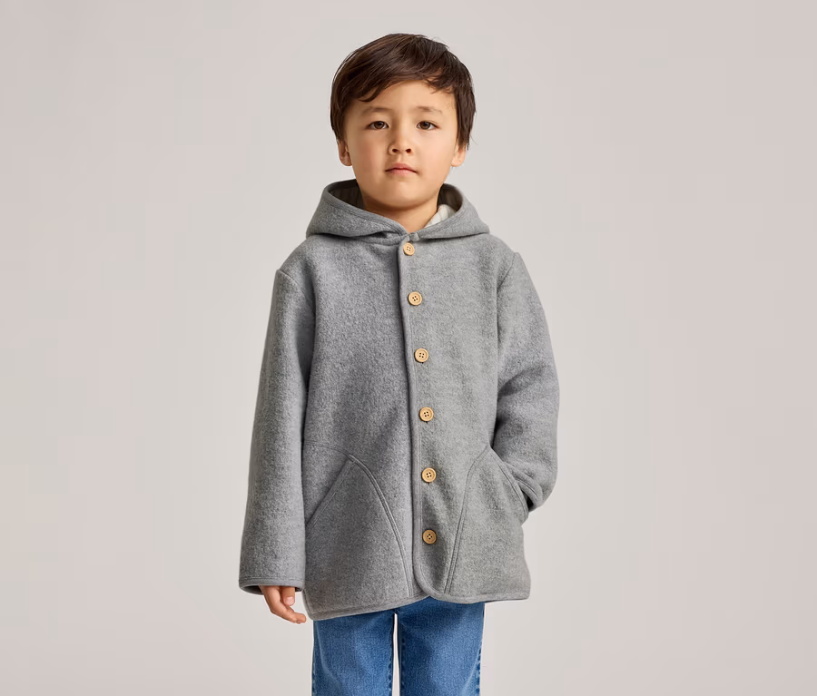 Un jeune enfant aux cheveux foncés se tient de face devant un fond clair, portant un manteau gris à capuche avec des boutons en bois et un jean bleu. Une main est dans l'une des deux poches avant.