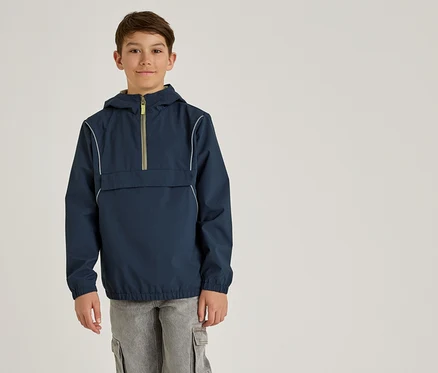 Un garçon porte un anorak bleu marine à capuche et un pantalon cargo gris sur un fond uni.