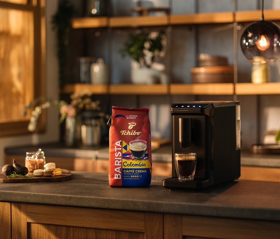 Sur un comptoir de cuisine, il y a une machine à café, une tasse de café et un paquet de BARISTA Origins Colombia en grains – 1 kg.