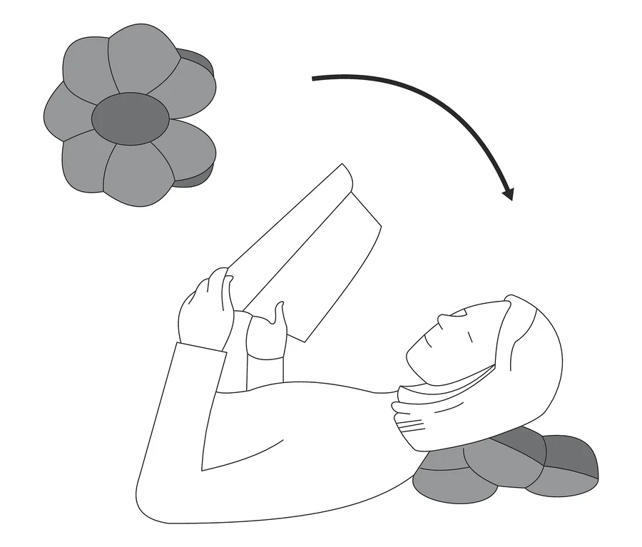 Femme allongée avec un coussin de relaxation multifonction sous la tête lisant un livre, fleur au-dessus.