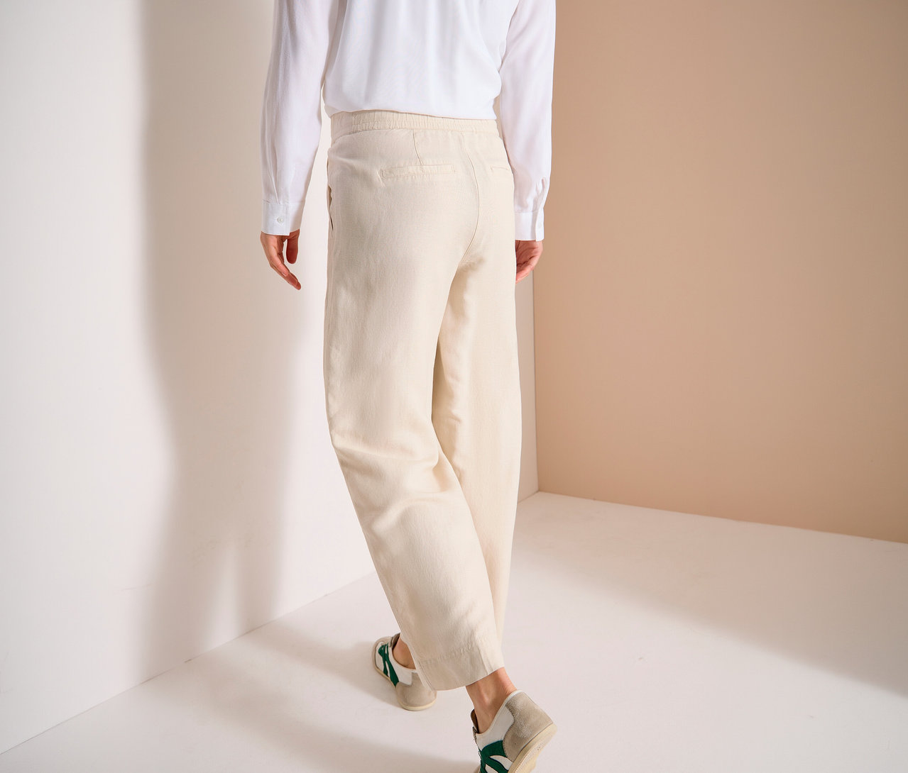 Personne vue de dos portant un pantalon beige, une chemise blanche et des baskets, debout contre un mur.