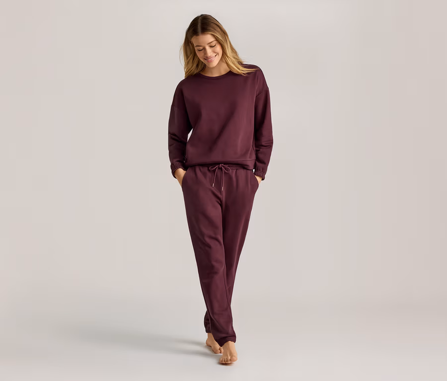 Une femme aux longs cheveux blonds porte un sweat-shirt bordeaux à manches longues et un pantalon de jogging. Elle marche pieds nus.