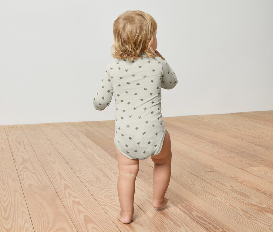 Un jeune enfant aux cheveux blonds porte un body vert clair à motif d'étoiles et marche sur un plancher en bois.