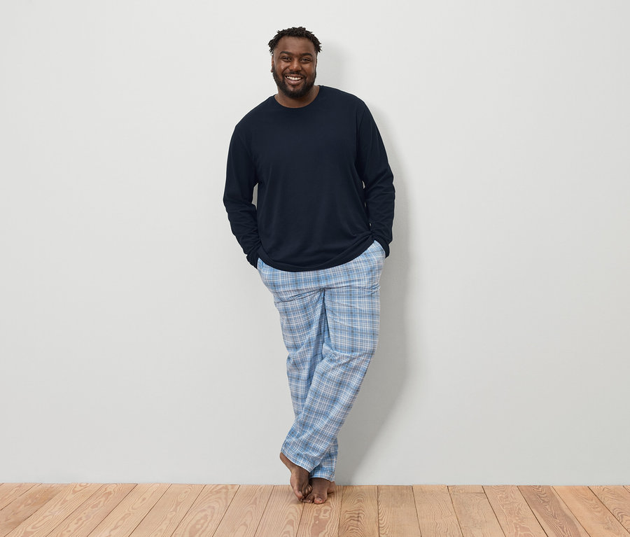 Un homme à la peau foncée avec une barbe, portant une chemise à manches longues bleu marine et un pantalon de pyjama à carreaux bleu et blanc, se tient pieds nus sur un plancher en bois devant un mur blanc.