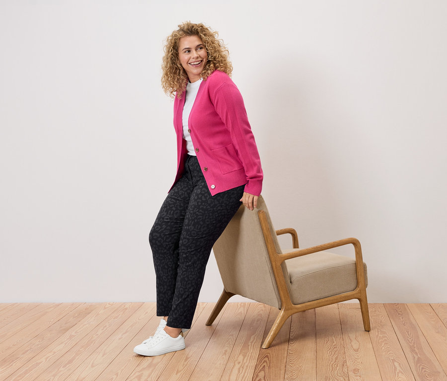 Femme aux cheveux bouclés, cardigan rose, pantalon à motifs et baskets blanches appuyée sur une chaise.