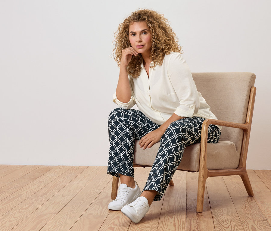 Une femme aux cheveux bouclés est assise dans un fauteuil avec un pantalon à motifs et des baskets blanches.