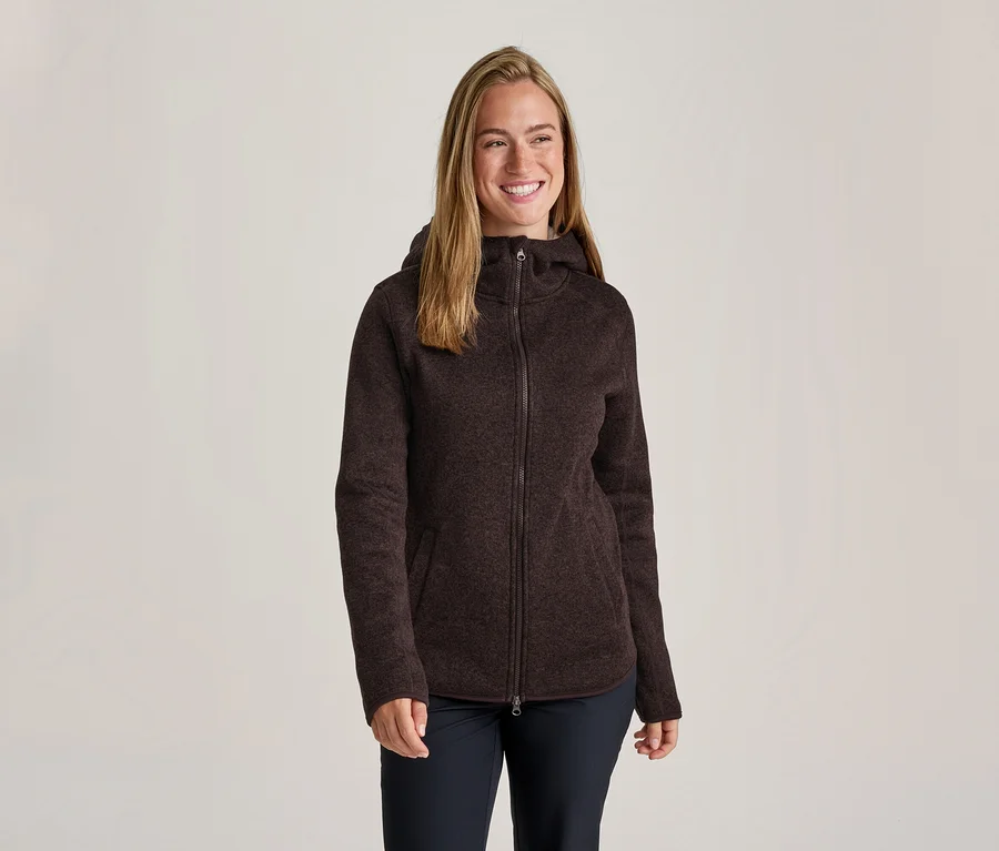 Femme souriante aux cheveux longs portant une veste polaire à capuche zippée texturée marron foncé et un pantalon foncé.