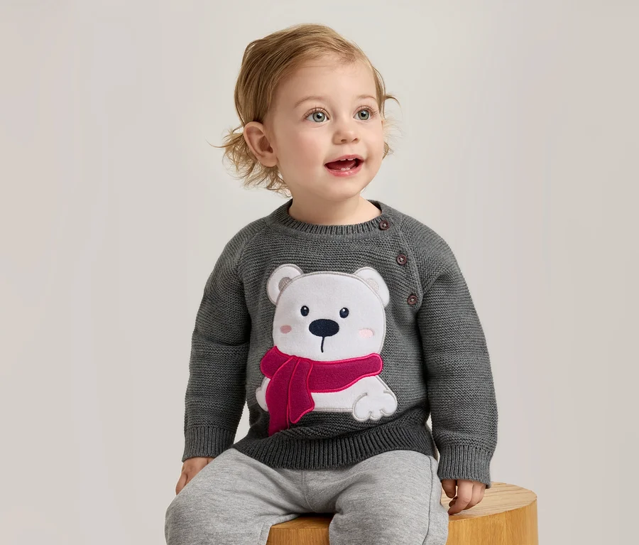 Un bébé aux boucles blondes est assis sur un tabouret en bois, portant un pull tricoté gris foncé avec une application d'ours polaire blanc, un foulard rose et des joues roses. Le bébé regarde en l'air, la bouche ouverte.