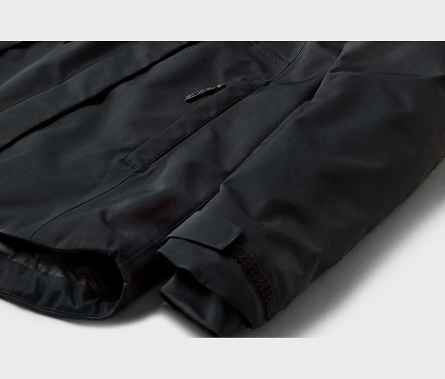 Gros plan d'une veste noire, montrant une poche poitrine zippée avec un logo sur la tirette et un poignet de manche ajustable avec fermeture auto-agrippante.