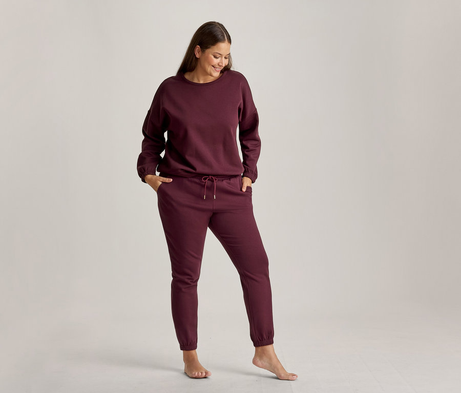 Une femme souriante regarde vers le bas, vêtue d'un ensemble de détente bordeaux composé d'un sweat-shirt et d'un pantalon de jogging. Ses mains sont dans les poches du pantalon et elle est pieds nus.