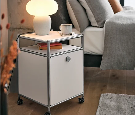 Lampe de table avec abat-jour en verre sur caisson en métal à roulettes « CN3 » avec abattant, blanc, à côté du lit.
