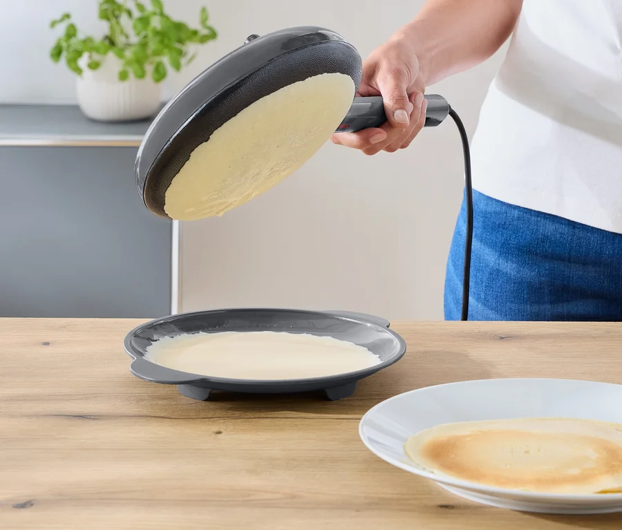 Une personne incline une poêle à frire électrique gris foncé au-dessus d'une assiette contenant le reste de la pâte. Une crêpe finie repose sur une assiette blanche sur un comptoir en bois.