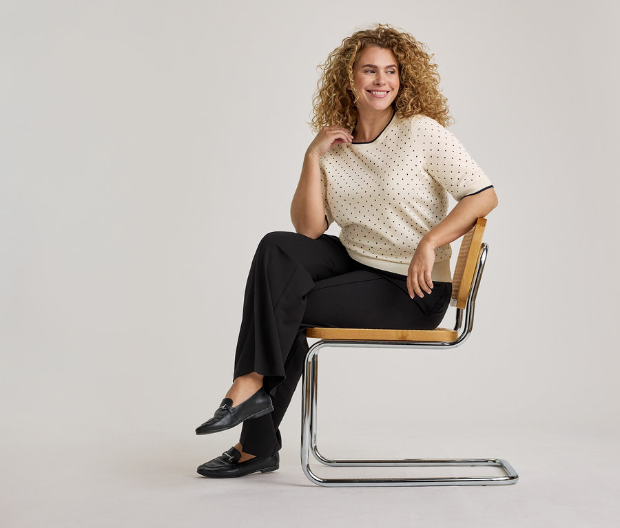 Une femme souriante aux cheveux bouclés est assise sur une chaise. Elle porte un haut crème à manches courtes avec des pois noirs et une bordure noire, un pantalon large noir et des mocassins noirs.