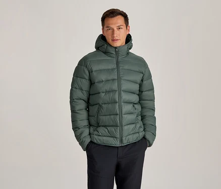 Homme portant une doudoune verte à capuche et un pantalon foncé, debout les mains dans les poches.